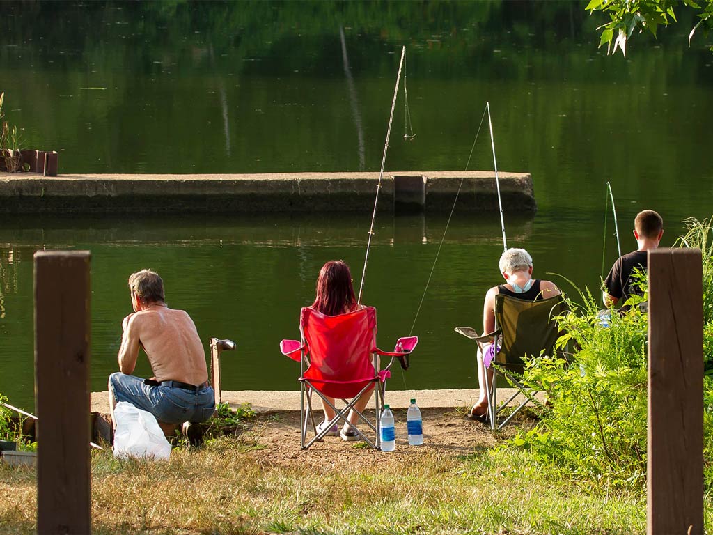 Four people sit in chairs by a calm lake, each holding fishing rods that evoke a serene angling scene. Two poles are propped up in holders, and lush greenery surrounds them. A wooden post is visible in the foreground.
