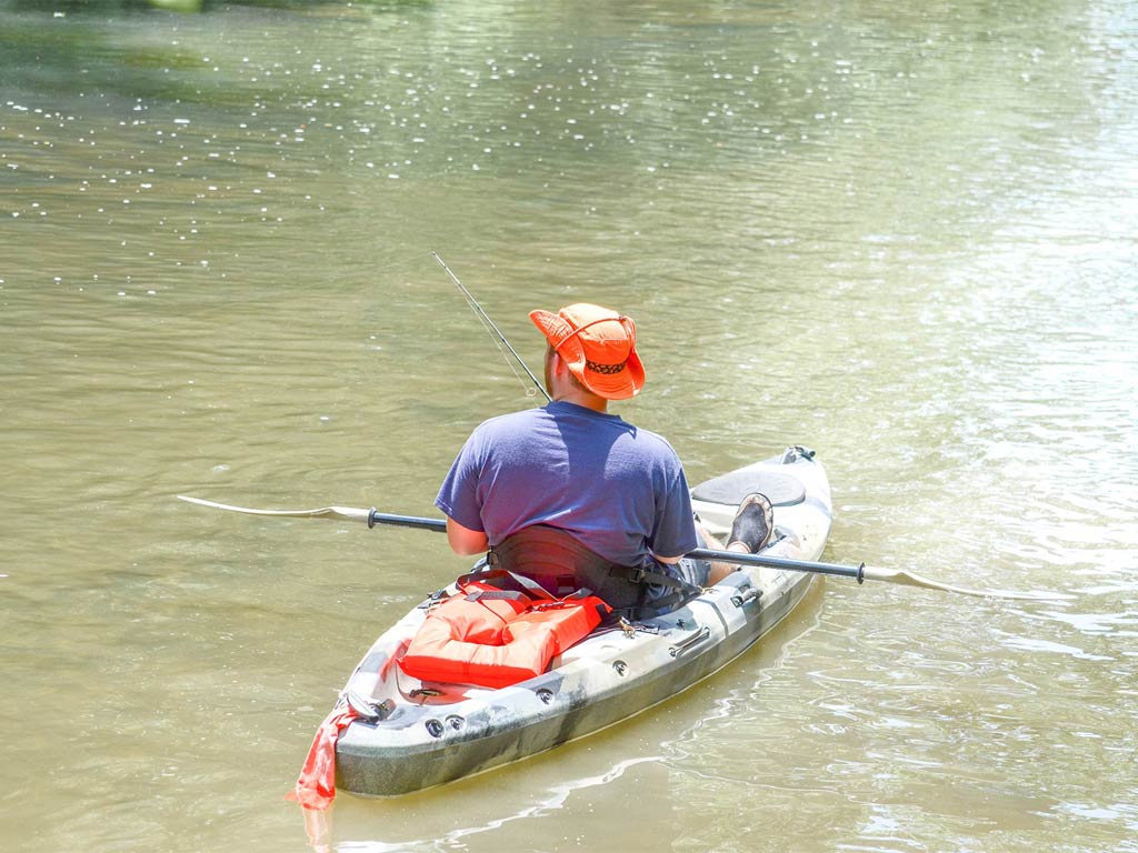 A person in a bright orange cap and blue shirt is kayaking and navigating peacefully with a fishing rod in hand and a red life jacket strapped to the kayak.