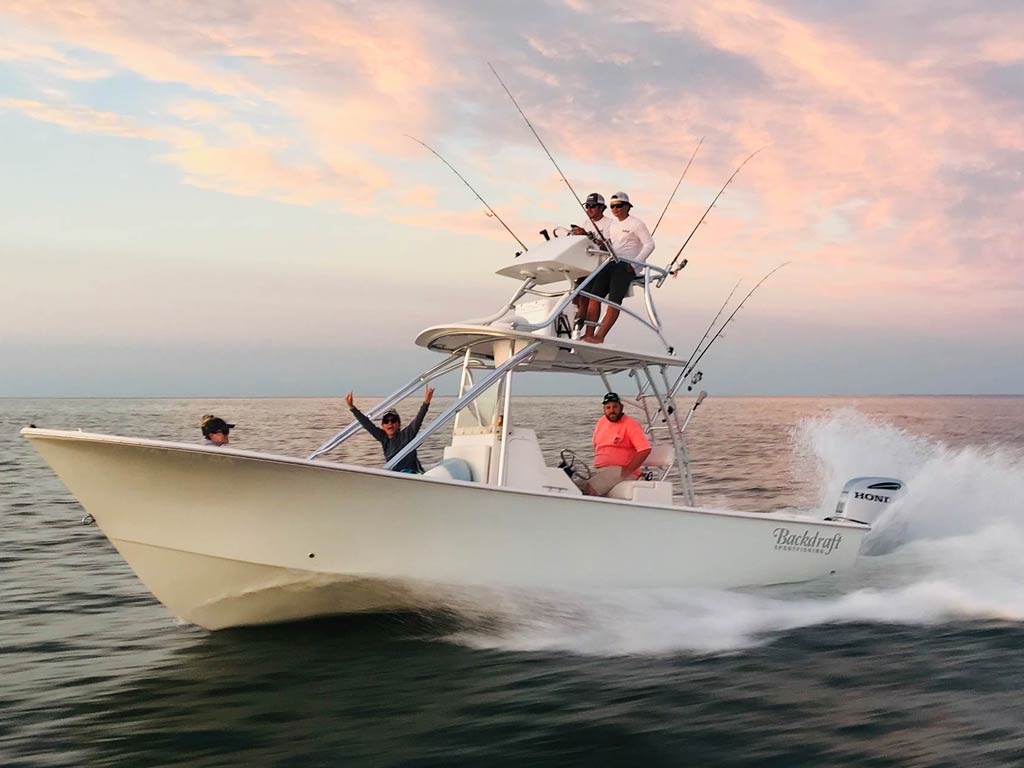 A white charter boat swiftly moves on the waters of Chesapeake Bay. Four people are aboard: one's seated at the helm, two stand on the platform above, and another waves from the side. The sky's painted with pink and blue hues, making it a perfect day for angling.