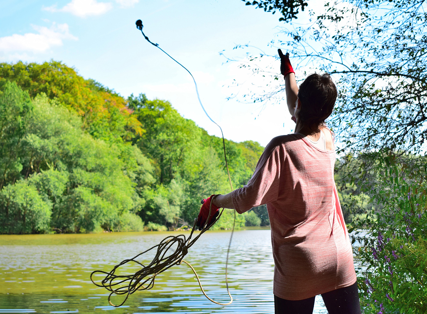 A person wearing gloves throws a rope with a Magnet attached into a Lake surrounded by Trees and Foliage.