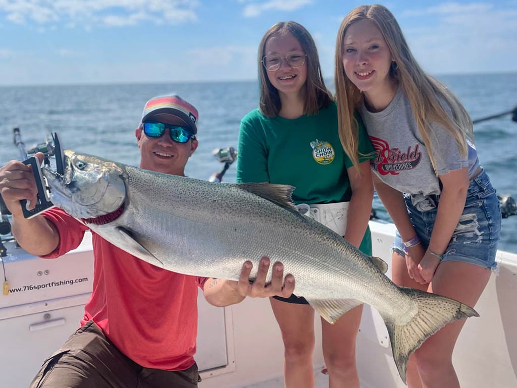 An angler in sunglasses and a hat smiling and kneeling on a charter fishing boat in Pulaski, NY, holding a huge Chinook Salmon, with two girls to the right of him leaning in to pose for the photo.