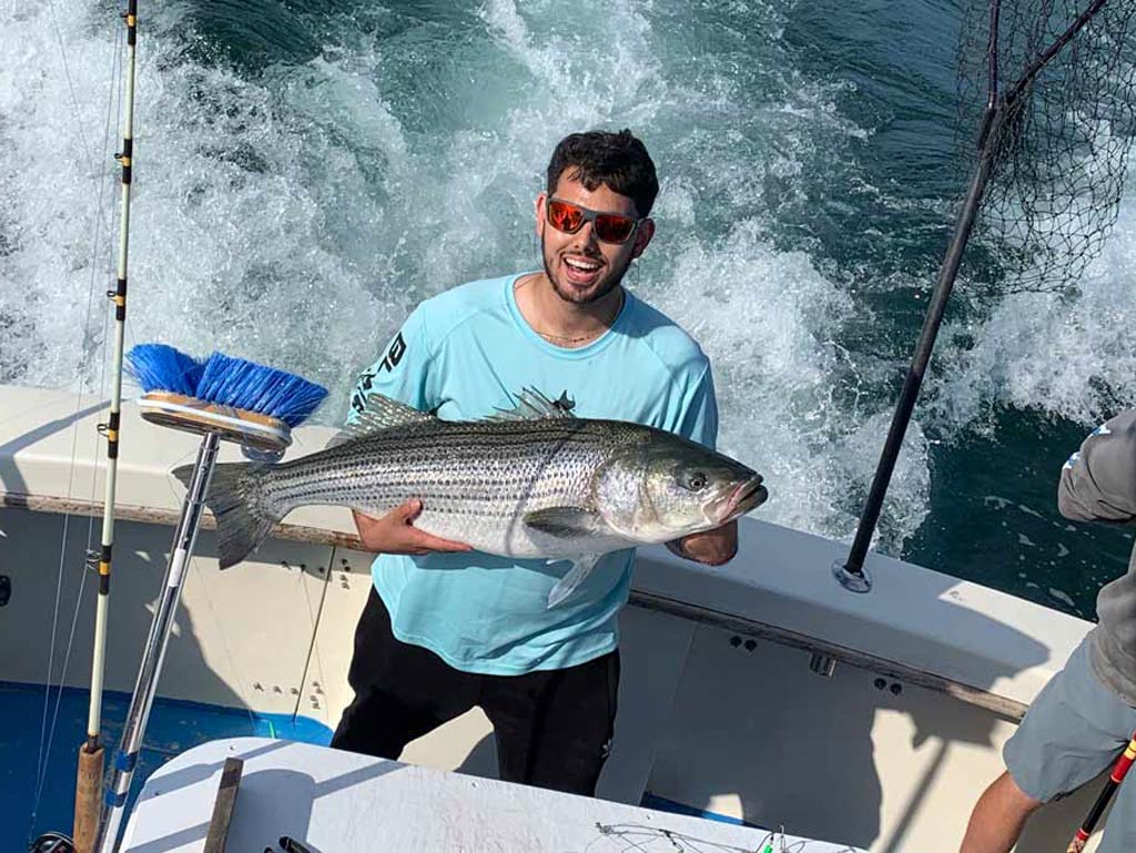 A smiling angler in sunglasses standing on the back deck of a charter boat, holding a sizeable Striped Bass he caught fishing in Montauk, New York, with the waters and the wake of the boat visible behind him.