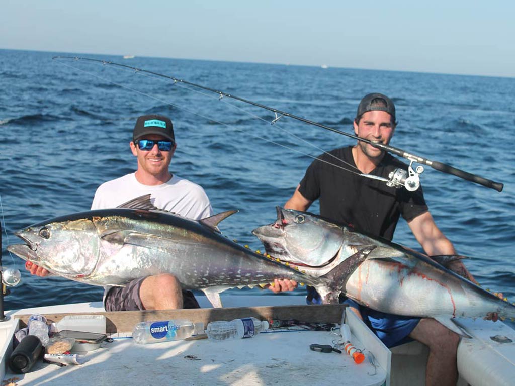 Two anglers posing for a photo on the deck of a boat, each holding a sizeable Bluefin Tuna, with the angler on the right biting down on his fishing rod and the ocean extending behind them.