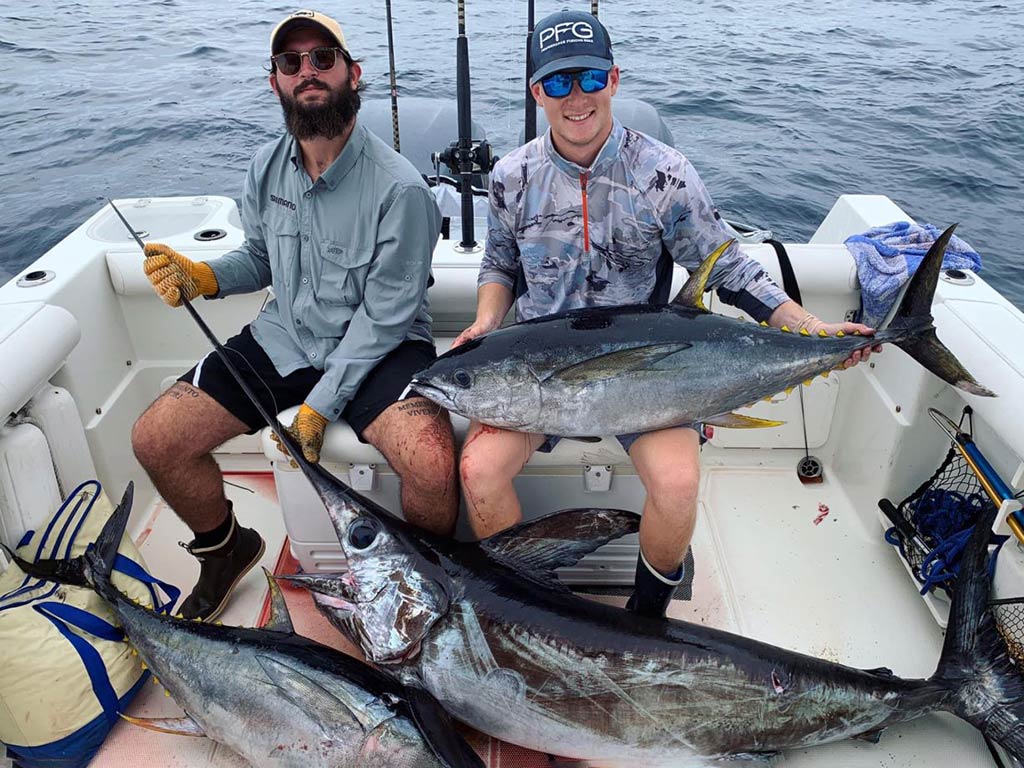 Two men sit on a boar in the Atlantic Ocean, with a Swordfish and Tuna in front of them and another Tuna across one of their knees.