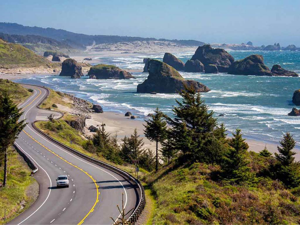 A view of Cannon Beach in Oregon, with a shoreline road to the left of the photo and the ocean to the right, with several rock formations sticking out of the water.