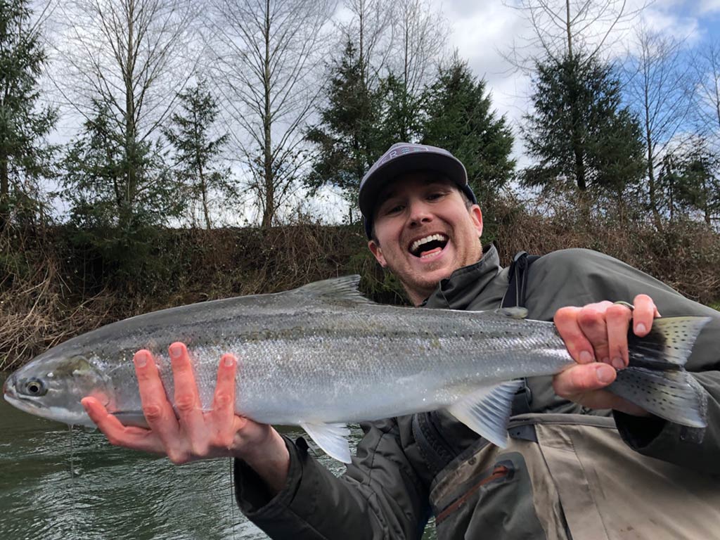 A man in a hat smiling and leaning back while holding a sizeable Steelhead he caught fishing in Oregon, with the river and shoreline foliage behind him.