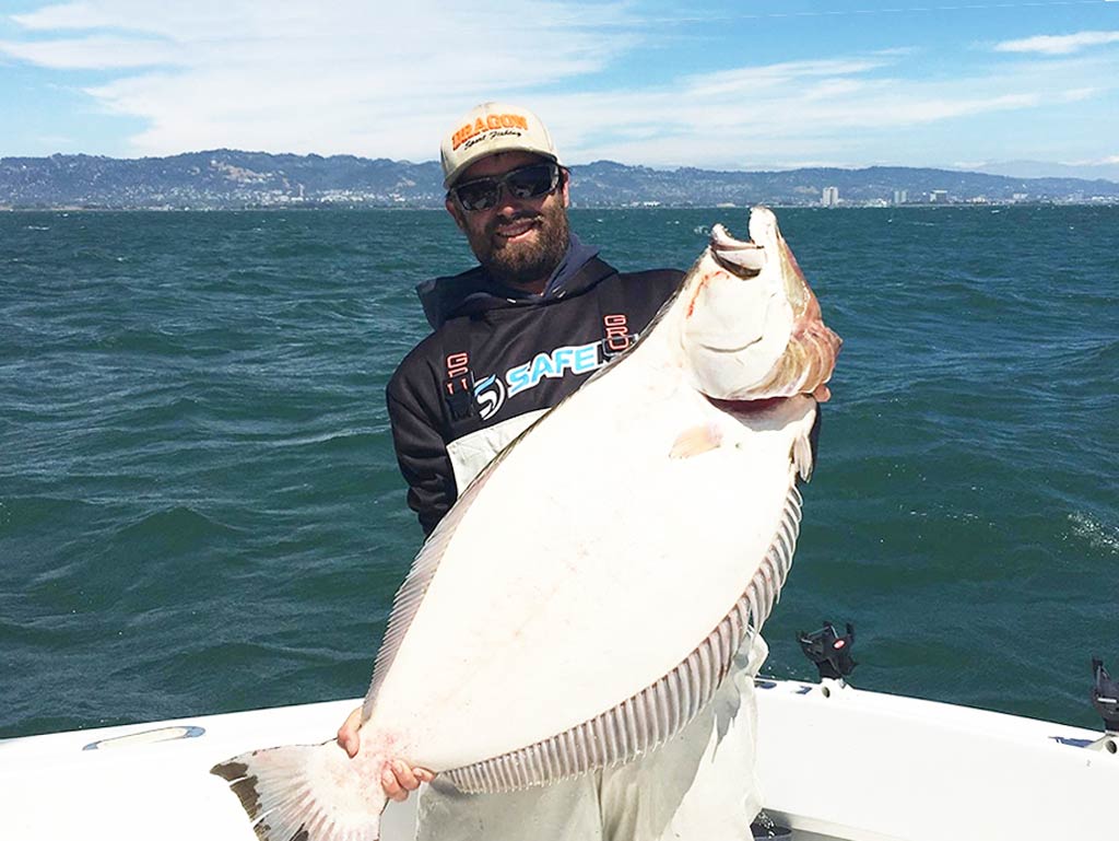 A man in sunglasses and a hat smiling and holding a large Halibut he caught fishing on an Oregon charter boat, with the waters and the shore visible in the distance.