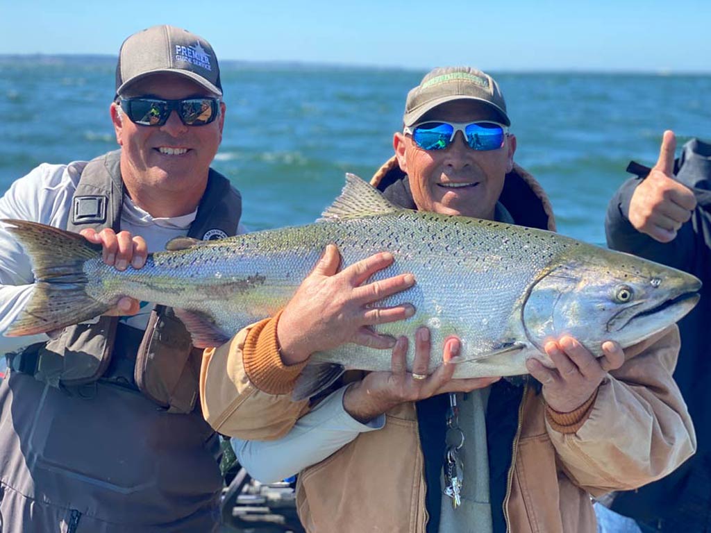 Two men in sunglasses and hats posing with a huge Salmon they reeled in fishing in Oregon, holding it at chest height, with the ocean behind them.