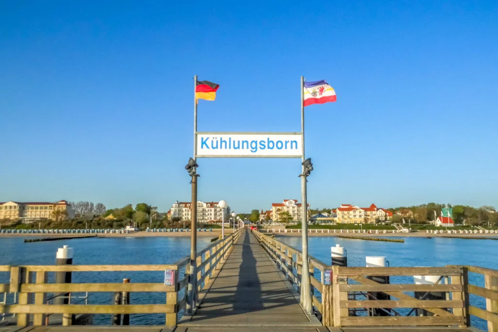 Eine h&ouml;lzerne Seebr&uuml;cke in K&uuml;hlungsborn ragt &uuml;ber das Wasser - wie geschaffen f&uuml;r Angeln an der Ostsee. Dar&uuml;ber wehen deutsche und regionale Flaggen, w&auml;hrend im Hintergrund Geb&auml;ude unter einem klaren blauen Himmel stehen. Angler k&ouml;nnen hier oft Arten wie Dorsch, Flunder, Scholle, Hering und gelegentlich auch Hornhecht oder Meerforelle fangen. Wenn Sie von der Seebr&uuml;cke aus angeln wollen, brauchen Sie nur ein paar Dinge: eine f&uuml;r die Ostsee geeignete Angelrute (in der Regel sechs bis neun Fu&szlig;), K&ouml;der wie W&uuml;rmer oder Kunstk&ouml;der, je nach Zielfischart, und eine g&uuml;ltige Angelerlaubnis, die nach den &ouml;rtlichen Vorschriften erforderlich ist. Verbringen Sie zwei bis drei Stunden am fr&uuml;hen Morgen oder am Abend, um die besten Chancen auf einen erfolgreichen Fang zu haben!