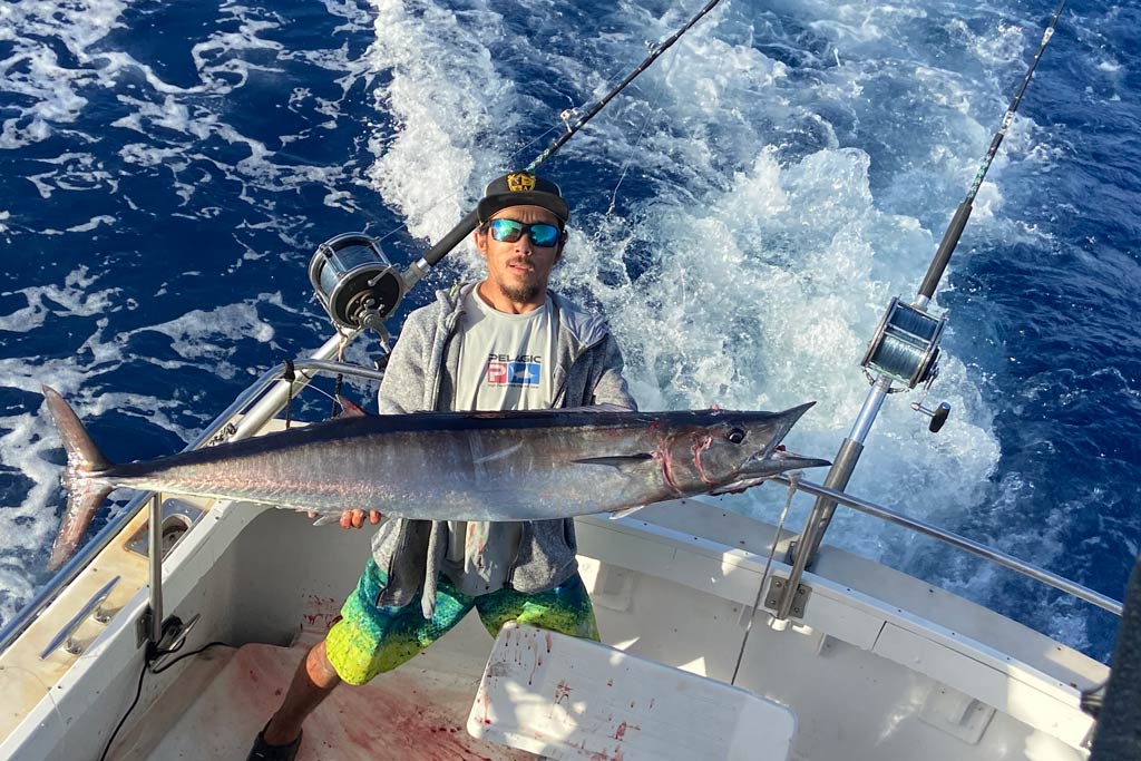 A fisherman in a cap and sunglasses standing in the corner of a boat, holding a big Wahoo with blue water in the background