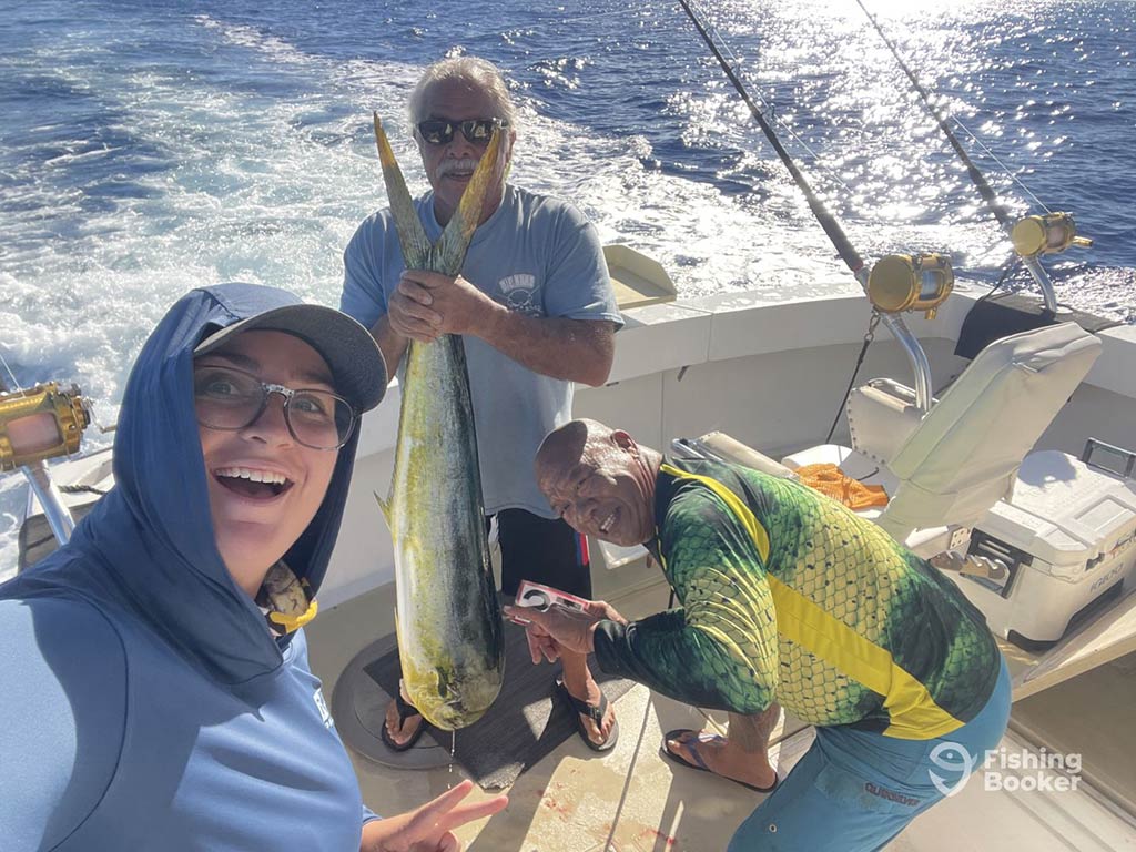 Three anglers celebrating on an offshore fishing boat, as the person in the middle holds a sizeable Mahi Mahi with the wake of the boat visible behind them.
