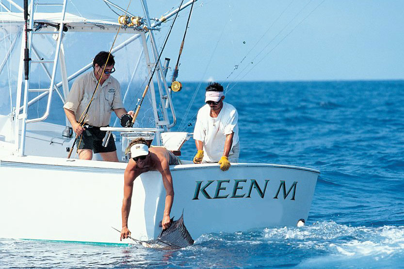 Three anglers aboard a boat, with one holding a Sailfish in the water by the bill and another holding a fly fishing line