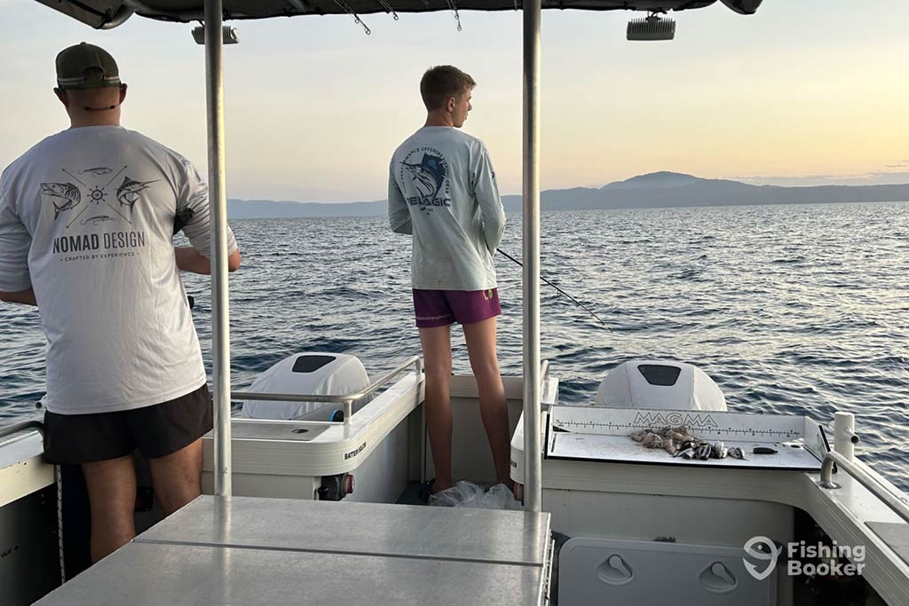 A view from behind of two anglers casting their lines from the deck of a fishing boat in Port Douglas, QLD, at sunset on some calm waters
