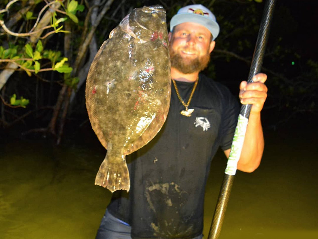 A closeup of a man holding a Flounder in one hand and a gig in the other while wading at night.