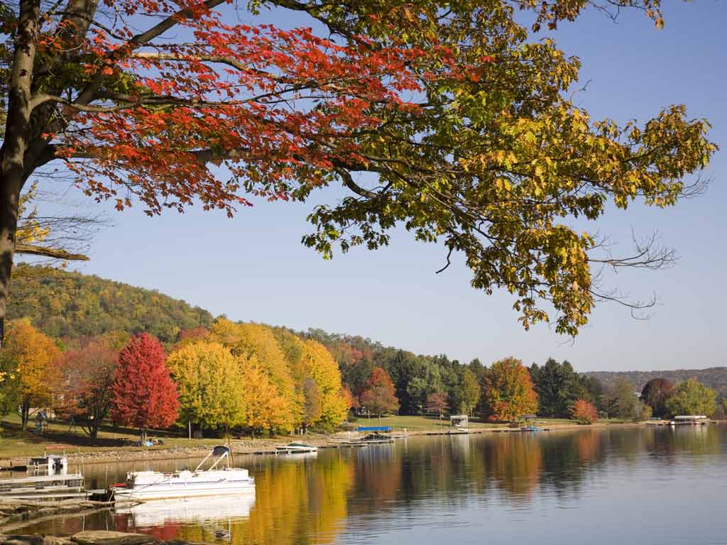 A photo of Deep Creek Lake in fall, with some boats in the water near the shore and colorful trees in the foreground and the background.