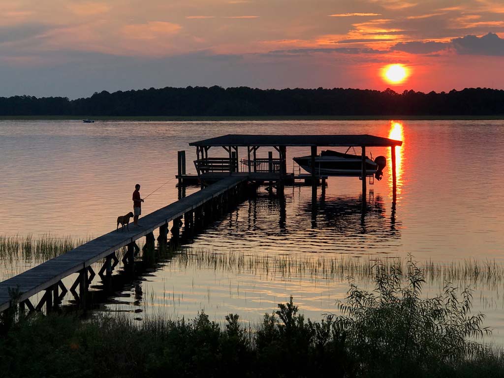 A man with a dog fishing from a pier at sunset.