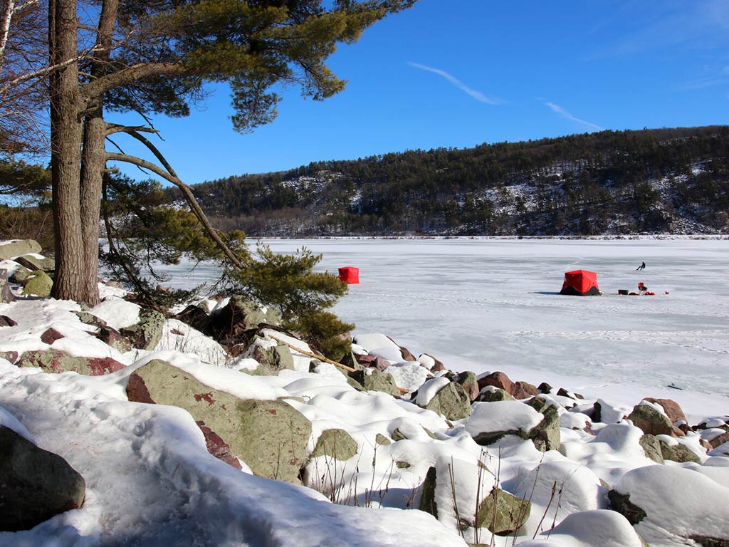 A snowy Wisconsin landscape features a frozen lake with two red ice fishing tents on the ice. The rocky, snow-covered shoreline frames a tree in the foreground, while forested hills rise in the background under a clear, blue sky