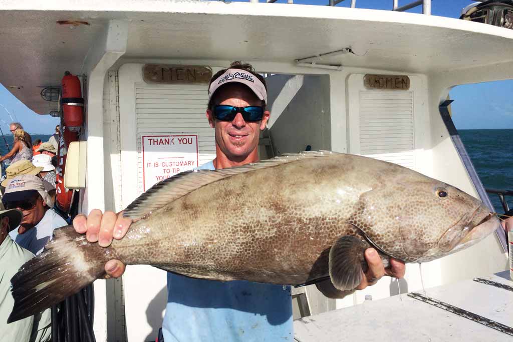 An angler in a cap and sunglasses holding a big Grouper on a party boat