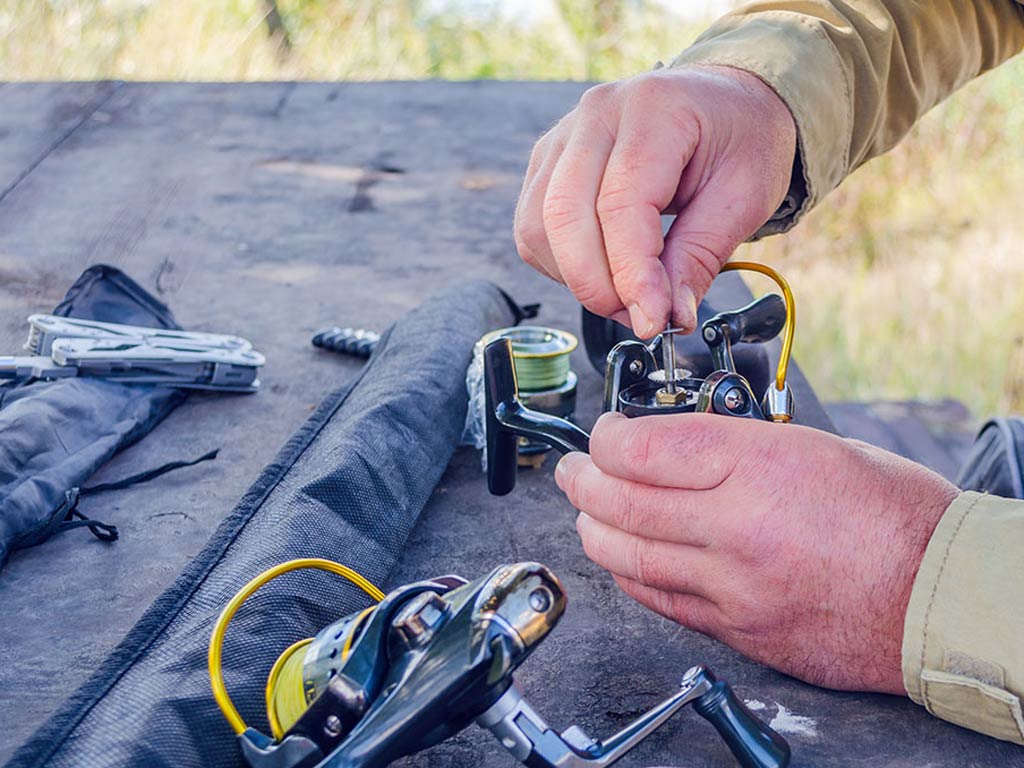 A man's hands screws into a fishing reel in the open air. The reel is made from aluminum which is the subject of the new tariffs in the US.