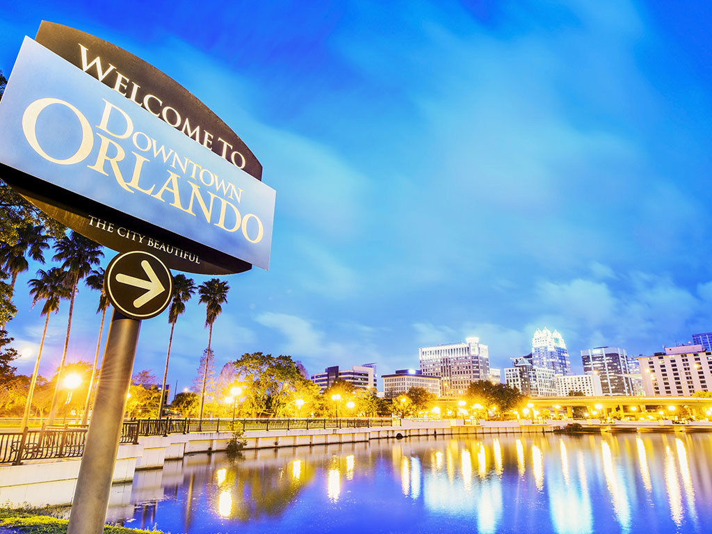 A view of the Orlando skyline across the water at night with a sifgn saying "Welcome to Downtown Orlando" in the left