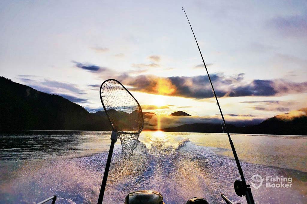 A view towards the back of a fishing boat with a lone trolling rod and fishing net perched towards the water and the sun setting in the distance in Ucluelet