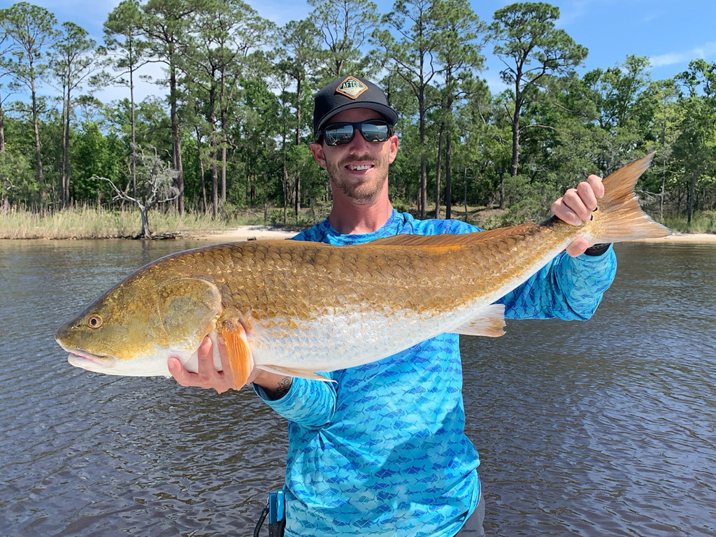 A beautiful photo of a proud angler holding Bull Redfish with both hands while standing on a charter fishing boat and posing against shoreline and trees in the background