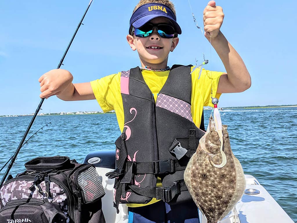A smiling boy standing on a boat, holding a Flounder he caught with a soft plastic lure