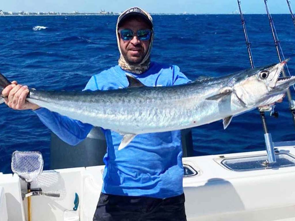 A fisherman holding a king Mackerel, standing on the side of a boat in a sun cap and sunglasses