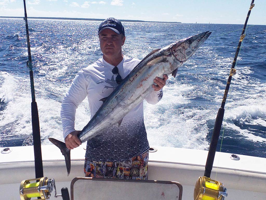 A fisherman standing on a fishing boat, between two rods, holding a Wahoo