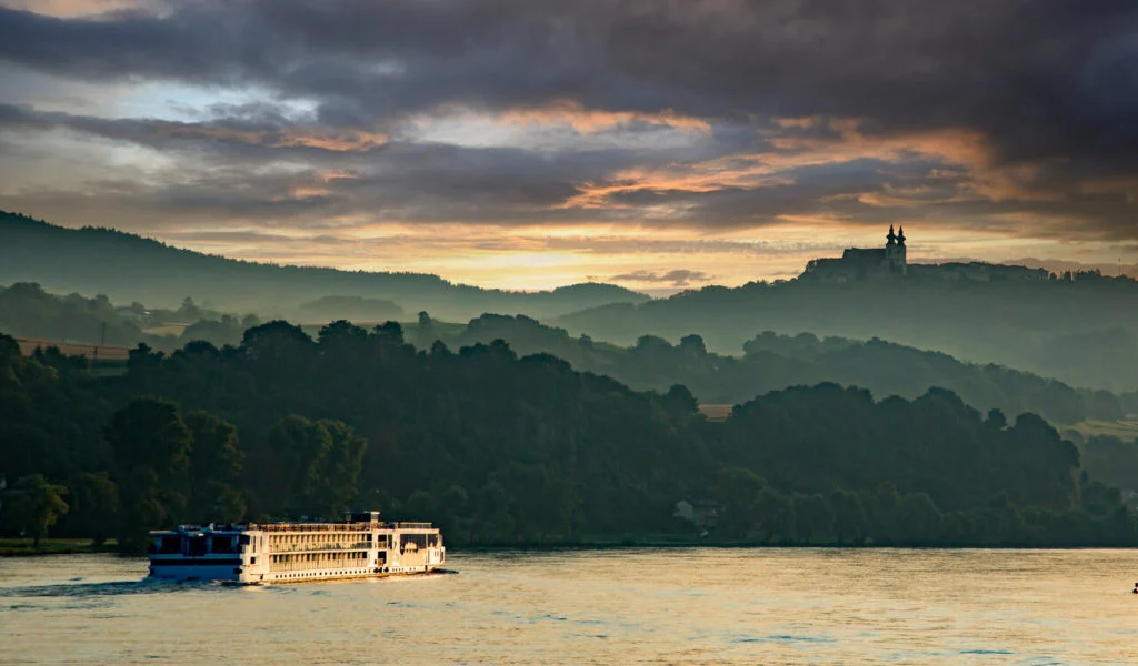Ein Flusskreuzfahrtschiff gleitet bei Sonnenuntergang &uuml;ber einen ruhigen Fluss und eignet sich damit perfekt f&uuml;r Angeln im deutschen Raum. Auf beiden Seiten erheben sich baumbewachsene H&uuml;gel, und in der Ferne ragt ein Geb&auml;ude in Form einer Silhouette auf. Angler in Deutschland hoffen, an solch friedlichen Abenden Arten wie Karpfen, Hecht, Barsch, Zander, Bachforelle oder Europ&auml;ischen Wels zu fangen. Ob man nun auf einen gro&szlig;en Karpfen hofft oder einfach nur die Landschaft genie&szlig;t, w&auml;hrend man in der D&auml;mmerung zwei oder drei Stunden lang angelt - es ist ein idealer Ort f&uuml;r Angelfreunde.