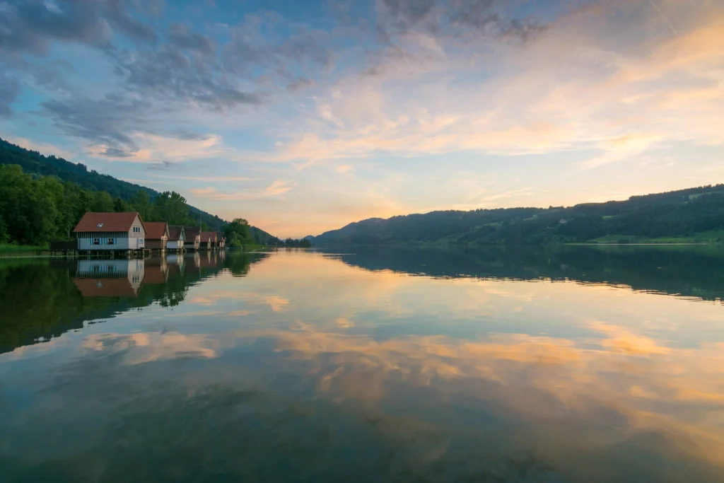 Ein ruhiger See, perfekt f&uuml;r Angeln im deutschen Raum, spiegelt Wolken und Himmel bei Sonnenuntergang. H&auml;user s&auml;umen die linke Uferlinie, w&auml;hrend sich im Hintergrund bewaldete H&uuml;gel erheben. Die friedliche Umgebung ist ideal, um Hechte, Barsche, Zander oder Karpfen zu fangen - egal, ob Sie lebende K&ouml;der oder K&ouml;der verwenden. Stellen Sie sich vor, wie die Sonne tief steht und Sie geduldig auf einen Biss einer Schleie oder vielleicht sogar eines seltenen Welses warten. Mit seinen ruhigen Gew&auml;ssern und der atemberaubenden Landschaft werden Sie keinen besseren Ort finden, um nach einem langen Tag zu entspannen.
