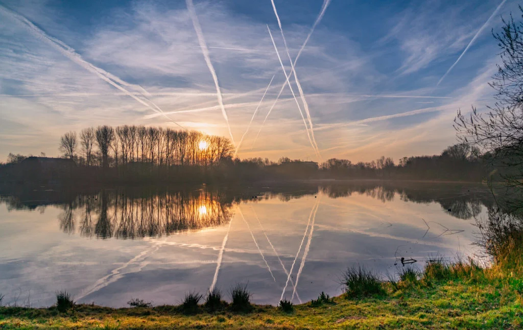 Sonnenaufgang &uuml;ber einem ruhigen See in Angeln im Deutschen Raum, mit B&auml;umen am Horizont und Flugzeugkondensstreifen, die sich im noch blauen Wasser spiegeln.