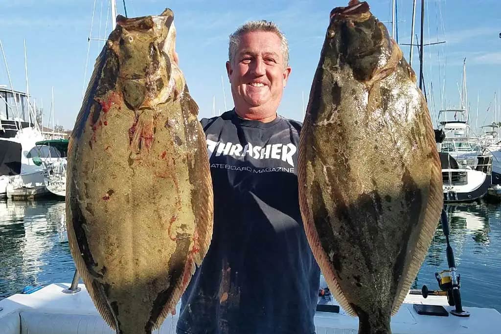 A smiling fisherman standing on a dock, holding two big California Halibut