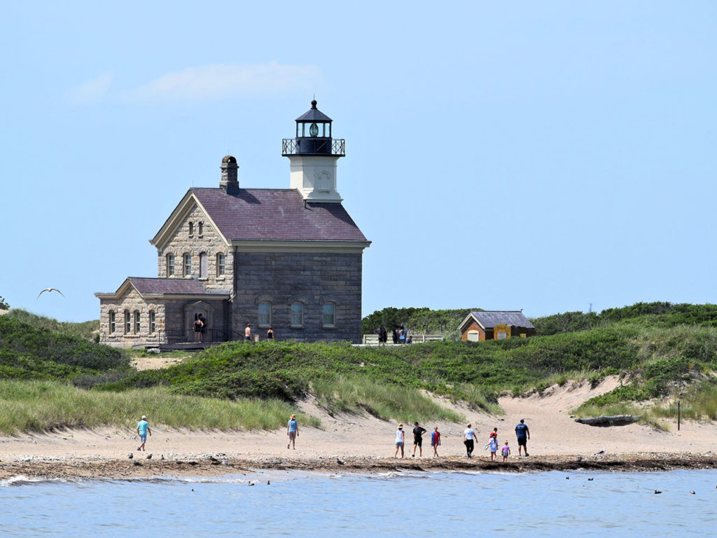 An old lighthouse overlooks a beach with people staring out to sea in Block Island, RI