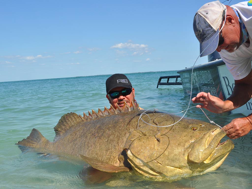 Two men proudly hold a large Goliath Grouper in the water by their boat in Clearwater, fresh from a thrilling deep-sea fishing adventure.