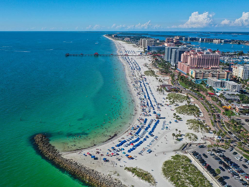 An aerial view of Clearwater Beach, Florida, showcases turquoise waters ideal for deep sea fishing. With its sandy beach and numerous umbrellas scattered about, it’s a picturesque setting. Nearby buildings add to the scene under a clear blue sky.