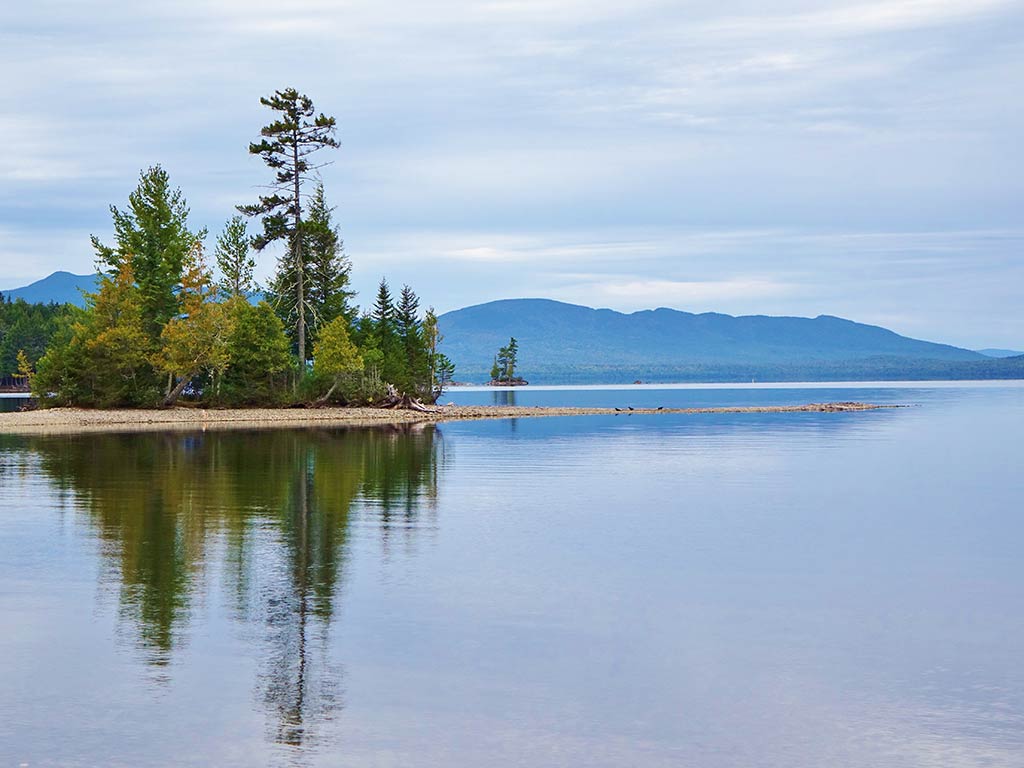 A photo of a calm Moosehead Lake on a cloudy day, with a couple of trees and mountain in the background.