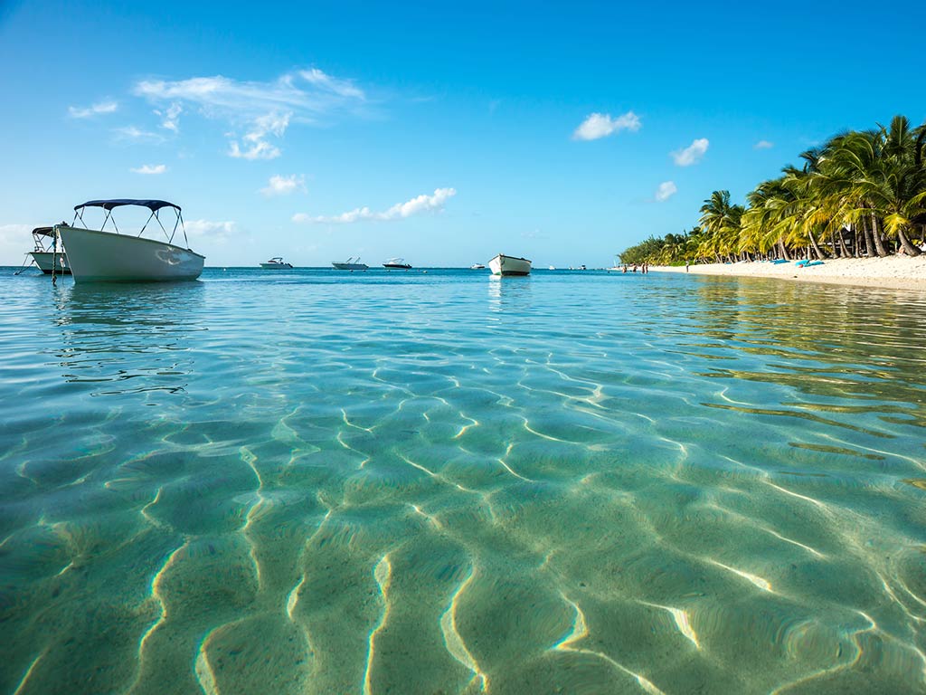 A photo featuring a charter boat peacefully standing still on the crystal clear water in the shallows next to a sandy beach