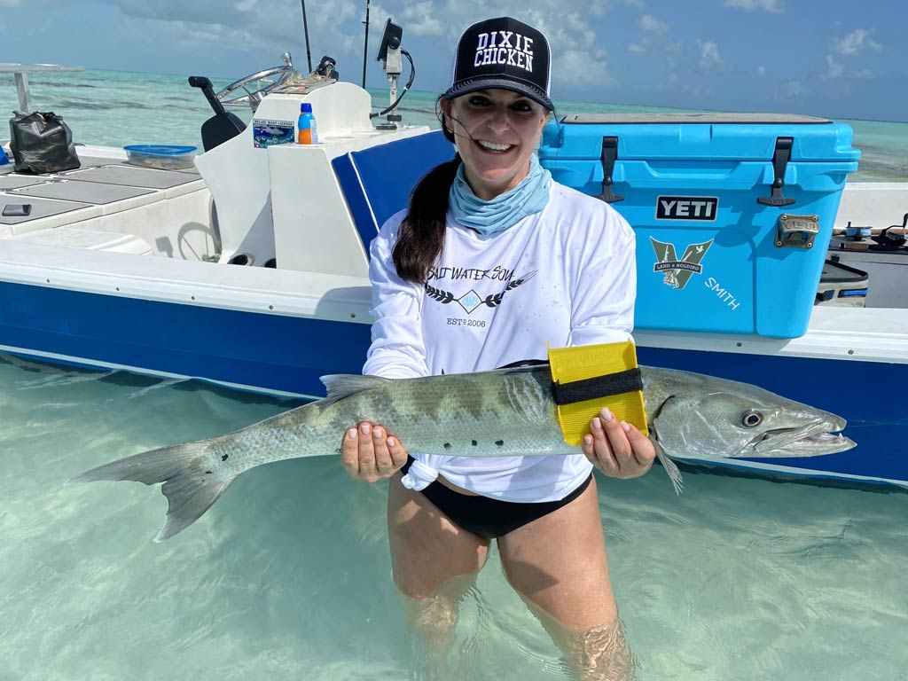 A smiling angler standing in the water in front of a charter boat and holding a freshly caught Barracuda while fishing in Belize