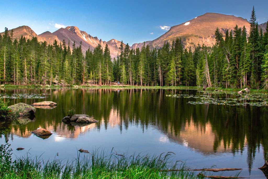 A view of Lake Estes in Estes Park, Colorado, with clear water mirroring evergreen trees surrounding it, and mountains in the background