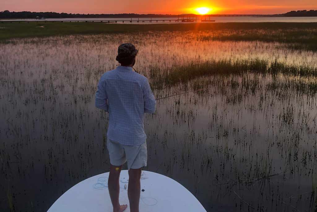 A fisherman standing on a boat, fishing, with sunset in front of him