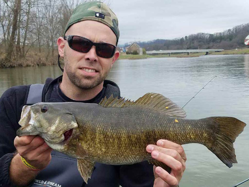 A fisherman in a cap put backwards and sunglasses, holding a Smallmouth Bass