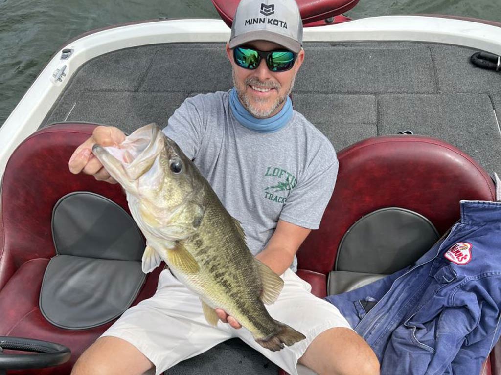 A photo featuring a middle-aged angler sitting on a charter boat and posing with a Largemouth Bass caught in Tennessee