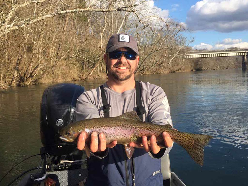 A smiling angler standing on a fishing boat, holding a Rainbow Trout