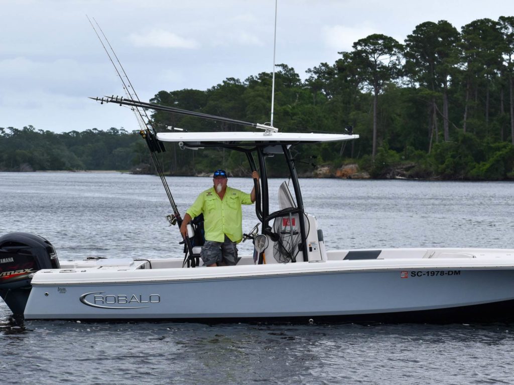 A view of a saltwater fishing guide in North Myrtle Beach, South Carolina, standing on a charter fishing boat with trees and water behind him.