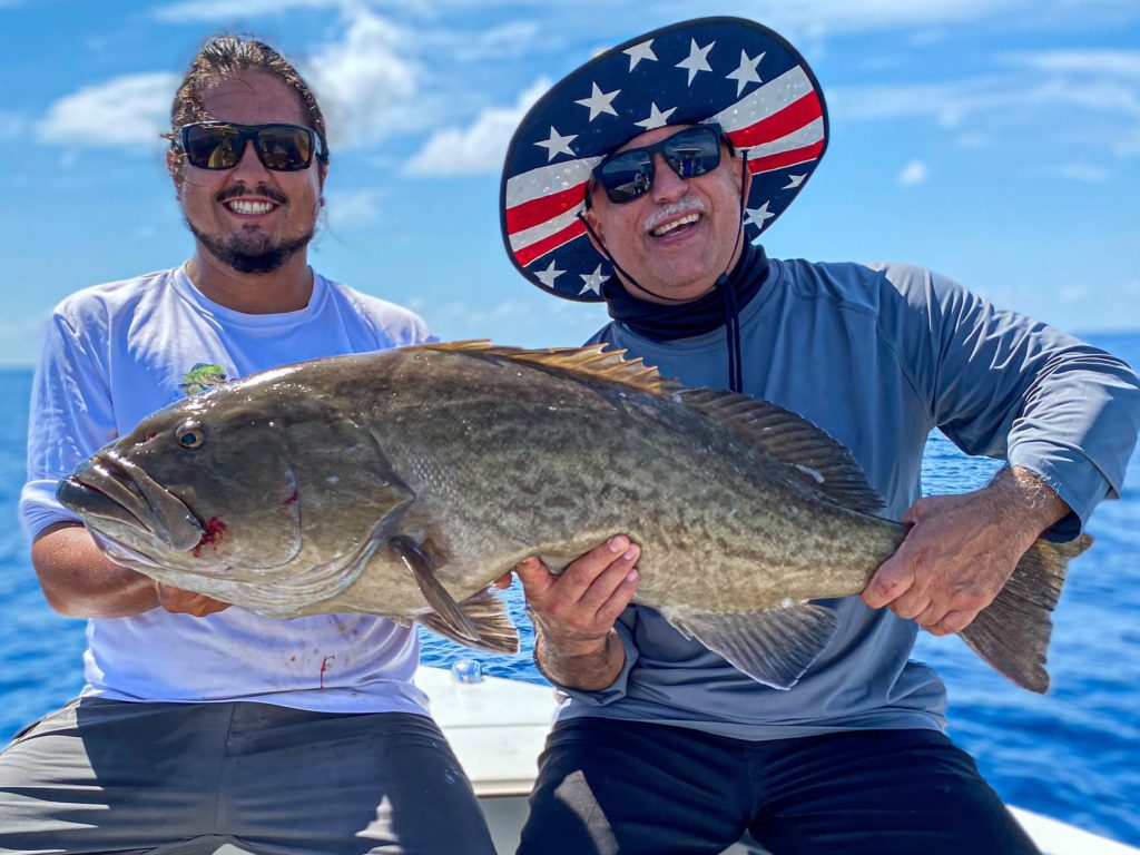 A picture showing two male anglers, both holding a freshly caught Grouper and wearing sunglasses on a fishing boat in Cape Canaveral, Florida