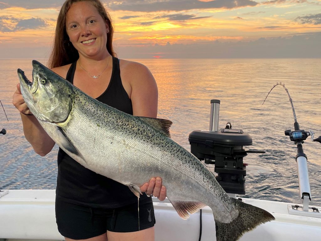 A smiling female angler holding a Salmon while standing on a charter fishing boat in Grand Haven, MI