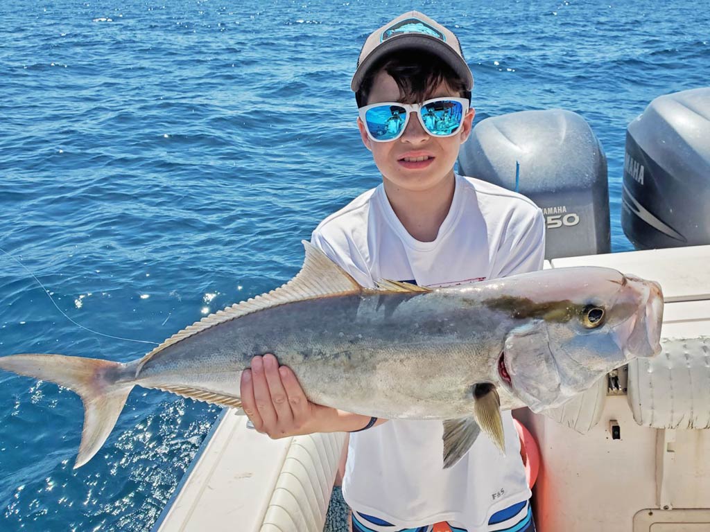 A photo of a kid holding smaller Amberjack catch while standing on a Tarpon Springs charter boat