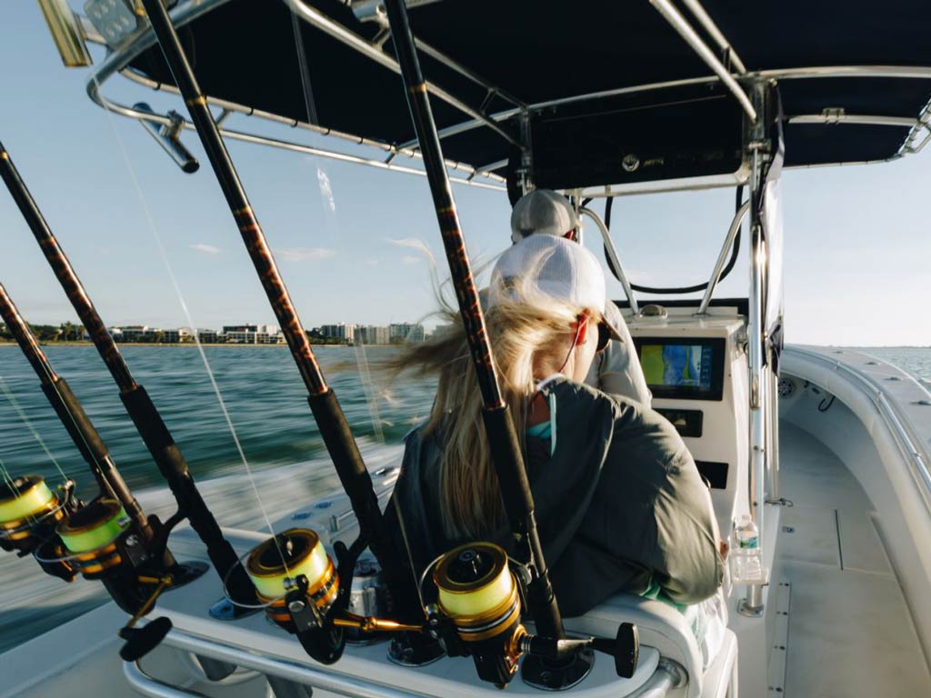 A photo of the rods, reels, and lines placed on a boat and a woman sitting in front of them while a captain is maneuvering the boat towards marina