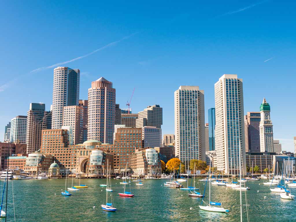 A photo of sailboats in the water off the coast of Boston, with the city's buildings visible in the background.